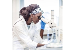 Image Woman in lab attire observing a sample under a microscope.